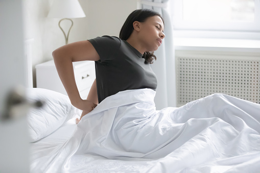 African mixed-race woman waking up sitting on bed feel low back pain after sleep caused by uncomfortable mattress, wrong position during night rest, premenstrual syndrome, violation of posture concept