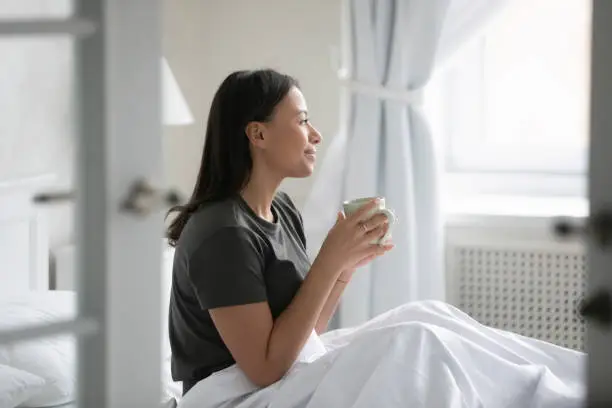 Modern apartment owner awakens enjoys good morning, start new day with cup of hot aromatic invigorating coffee, excellent mood, vacations concept. African woman wake up sit in bed holds mug drink tea