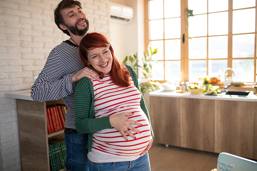 Lovely husband massaging his pregnant wife, while she holds hand on her stomach. They are standing in bright kitchen at home