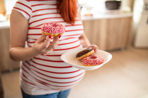 Midsection of pregnant woman standing at kitchen, holding plate and eating delicious doughnut at home
