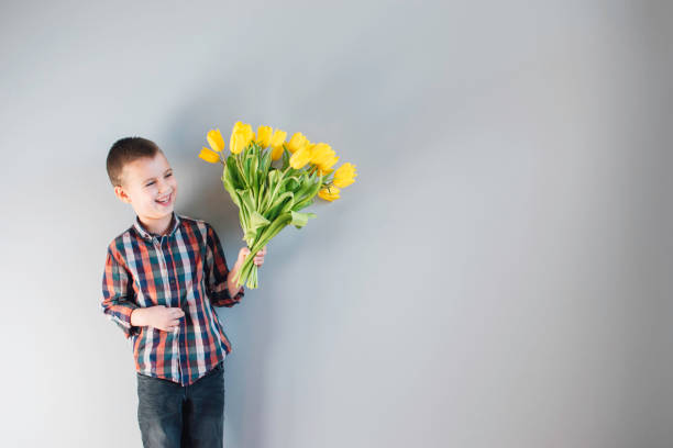 Cheerful little boy in plaid shirt holding big bouquet of yellow tulips on the grey background. Copy space.