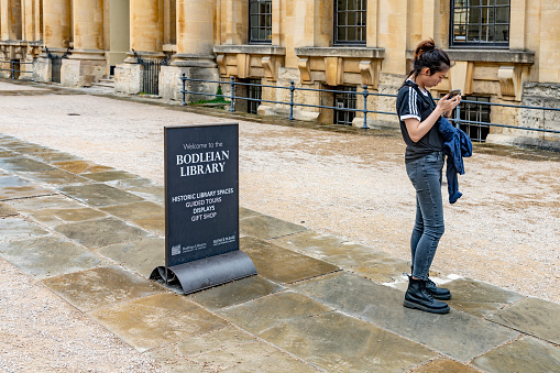 A young woman checking her phone outside the Bodleian Library and All Souls College at the university of Oxford. Oxford, Oxfordshire, England, UK