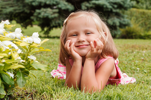 Little blonde girl in a pink dress lies on the grass with her head in her hands near the flowers and looks at the camera