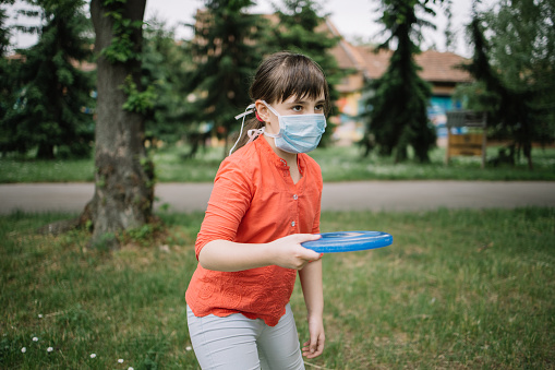 Side view of child holding flying disk and breathing through medical mask. Nice girl playing outside in front of green trees during the coronavirus pandemic