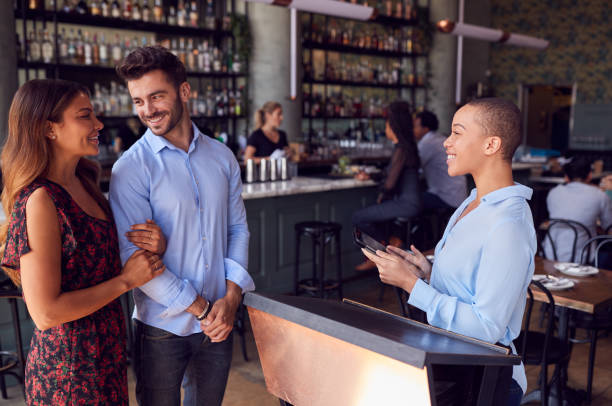 Couple Being Greeted By Maitre D Using Digital Tablet As They Arrive At Restaurant