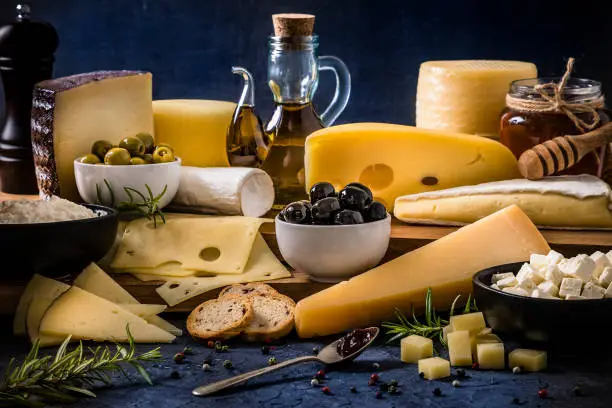 Front view of various types of cheese like Emmental cheese, cottage cheese, manchego cheese, parmesan cheese, brie cheese, cured and semi-cured cheeses on a bluish tint background. The composition includes black and green olives, an olive oil bottle, rosemary, a honey jar with a honey dipper, a pepper shaker and pepper. Predominant colors are blue and yellow. Low key DSLR photo taken with Canon EOS 6D Mark II and Canon EF 24-105 mm f/4L