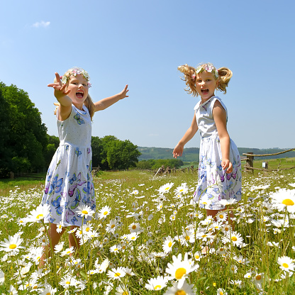 Twin sisters play together in a daisy \nfield and smile cheerfully at the camera. They\nboth jump up and down with laughter.