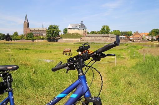 Kessel, Netherlands - May 21. 2020: View beyond handlebar of bicycle over green rural landscape on church and castle of medieval village (focus on handlebar)