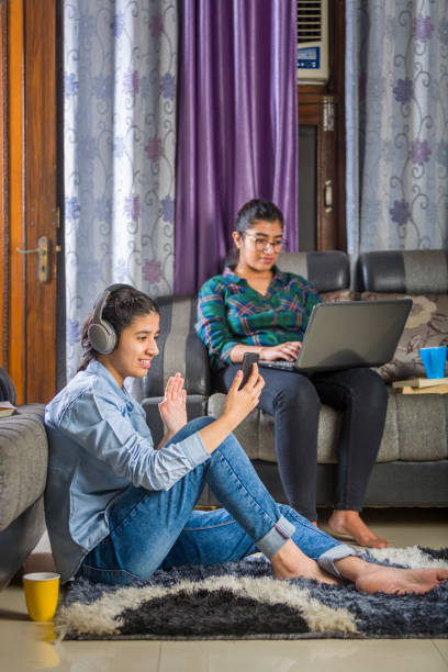 deux filles assises à la maison occupées dans un appel vidéo utilisant le smartphone, l’ordinateur portatif et le casque s’asseyant sur l’étage - indian girls audio photos et images de collection