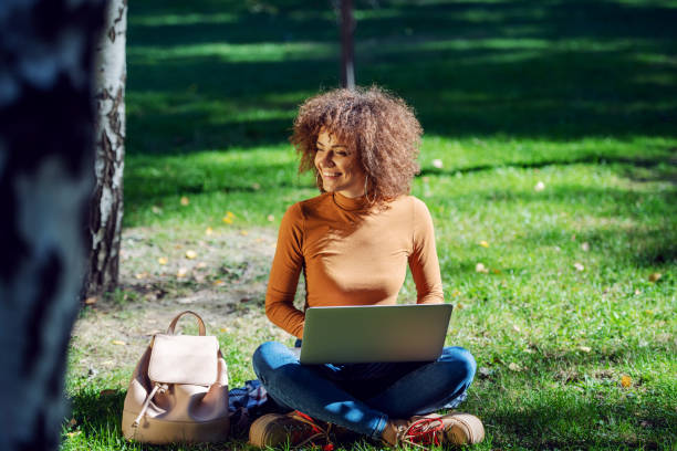 istock Young female student sitting on lawn in park and using laptop. 1226433387