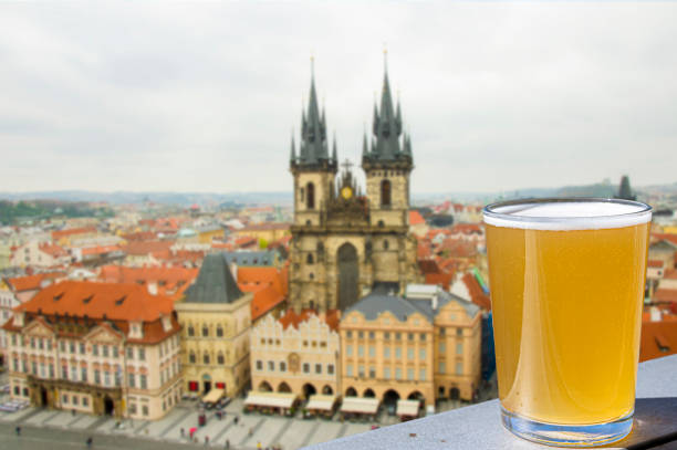 vista di un bicchiere di birra leggera con sfondo della piazza della città vecchia e della chiesa della madre di dio prima di tyn a praga, ceco - amber beer foto e immagini stock
