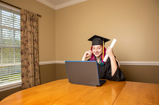 A high school girl graduating on her laptop and holding a rolled up diploma during the coronavirus pandemic.