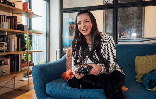 A shot of a mid adult Colombian woman sitting on the sofa, she is playing video games at her home in London, England.