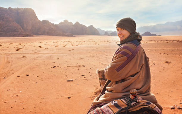 giovane donna in sella a cammello nel deserto, guardando indietro sopra la spalla, sorridendo. fa abbastanza freddo, quindi indossa il tradizionale cappotto beduino - bisht - e sciarpa per la testa, montagne di fondo - camel back foto e immagini stock