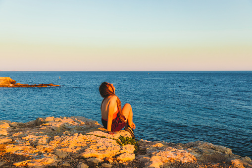 Happy woman with long hair and in swimsuit enjoying the picturesque colorful sunset from the cliff near the lighthouse and the  turquoise sea in French Riviera