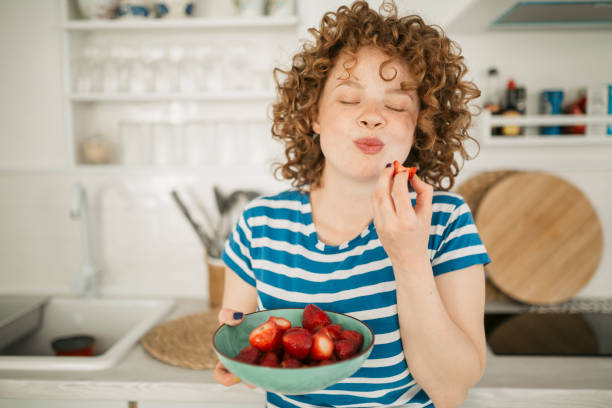 jeune femme de rousse gaie mangeant des fruits à la maison - petite faiblesse photos et images de collection