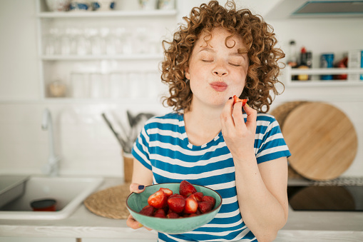 Beautiful young woman with red curly hair standing in the kitchen of her apartment, eating and enjoying a bowl of strawberries