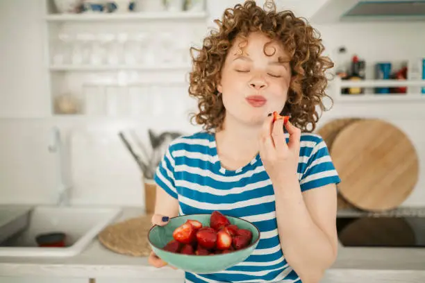 Beautiful young woman with red curly hair standing in the kitchen of her apartment, eating and enjoying a bowl of strawberries