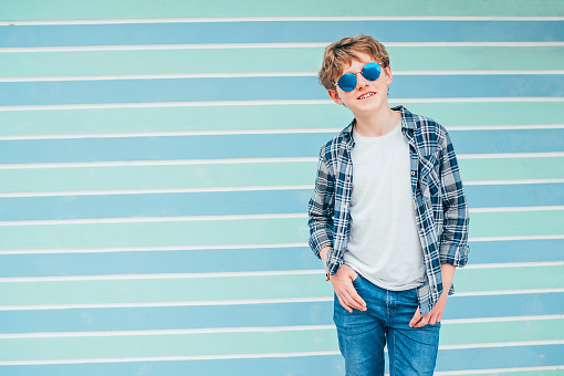 Caucasian blonde hair 12 year old teenager boy Fashion portrait dressed white t-shirt with checkered shirt in blue sunglasses with turquoise blue background wall background. Teens fashion concept.