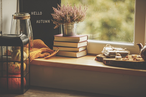 Side view of Hello Autumn sign placed against the window in day light with set of books, decorative flowers, orange scarf and lantern with decorative pumpkins.