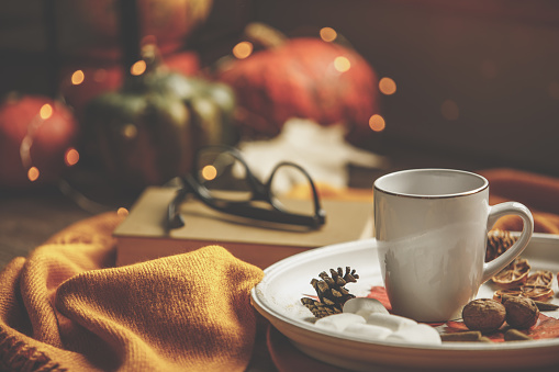 Front view of hot beverage  cup with almond treats and marshmallows placed on top of orange blanket with colorful decorative pumpkins and reading glasses in the background.