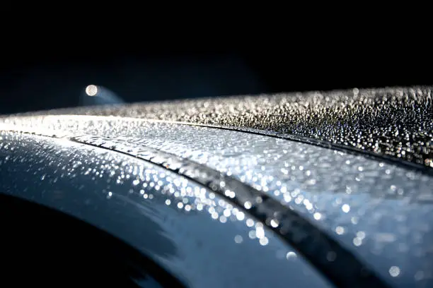 Evening rain with water drops on a white car with tinted windows and sunroof. Low-key, dark background, water droplets reflection, wet car.
