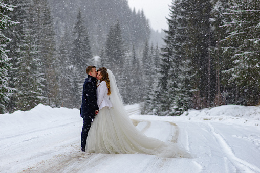 The groom kisses the bride against the backdrop of a snowy fir forest. Snowing. Winter wedding.