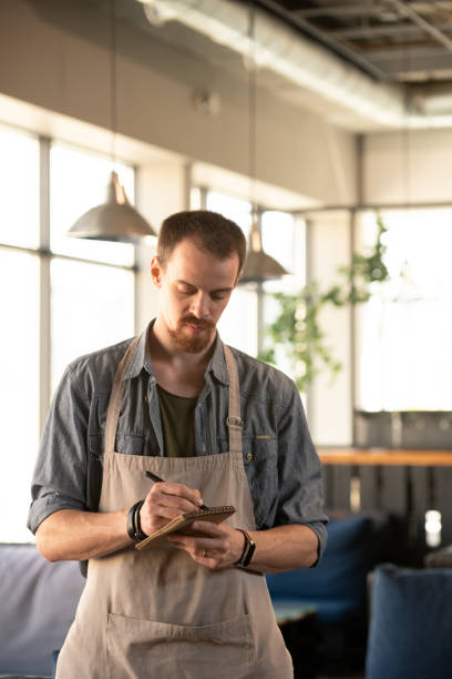 istock Young serious waiter in apron and shirt writing down order of client in notepad 1226530288