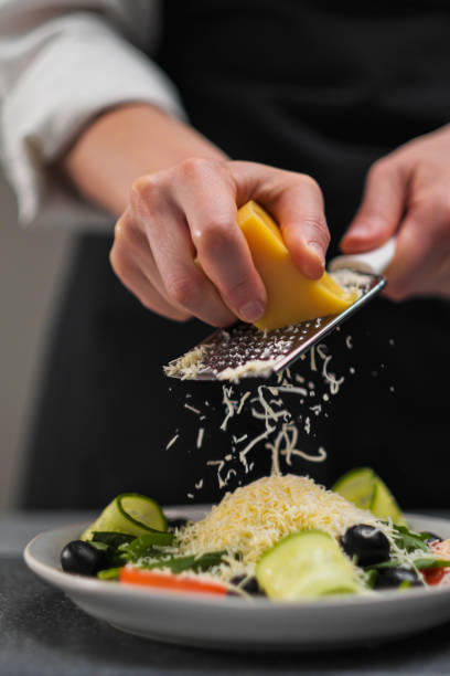 A female chef in a white uniform and a black apron in the restaurant kitchen. Cooking a salad. The cook rubs the parmesan cheese on a small grater. stock photo