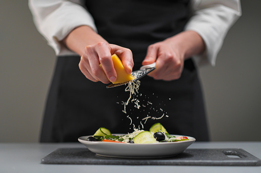 A female chef in a white uniform and a black apron in the restaurant kitchen. Cooking a salad of vegetables. The cook rubs the parmesan cheese on a small grater.