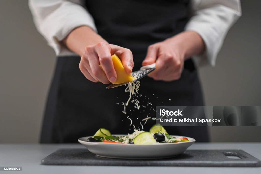 A female chef in a white uniform and a black apron in the restaurant kitchen. Cooking a salad. The cook rubs the parmesan cheese on a small grater. A female chef in a white uniform and a black apron in the restaurant kitchen. Cooking a salad of vegetables. The cook rubs the parmesan cheese on a small grater. Chef Stock Photo