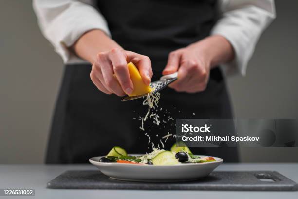A Female Chef In A White Uniform And A Black Apron In The Restaurant Kitchen Cooking A Salad The Cook Rubs The Parmesan Cheese On A Small Grater Stock Photo - Download Image Now