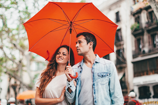 Happy smiling spanish Tourist Couple walking along the famous Ramblas Street in Barcelona under a red umbrella. Looking around happy and content. Tourist Couple Lifestyle Barcelona, Catalonia, Spain, Europe