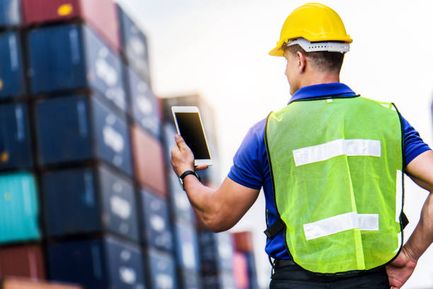 istock Young confident Caucasian man engineer using digital tablet and wearing yellow safety helmet and check for control loading containers box from Cargo freight ship for import and export, transport. 1226544994
