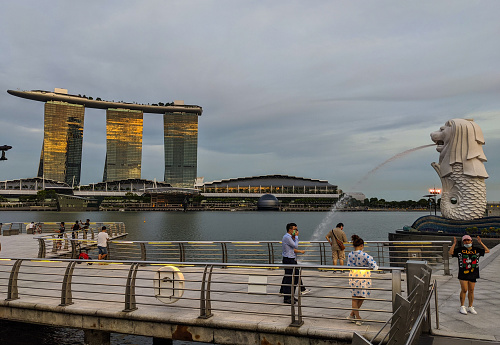 May 2020. Singapore. Several people seen in the shot taking picture near the famous monument in Merlion Park Singapore landmark and a major tourist attraction, located near One Fullerton, Singapore, near the Central Business District. In the background one can see the reflection of the sunset in Marina Bay Sands. The Merlion is a mythical creature with a lion's head and the body of a fish that is widely used as a mascot and national personification of Singapore. Some people are seen wearing masks while others don't wear masks during the COVID 19 / Coronavirus pandemic.
