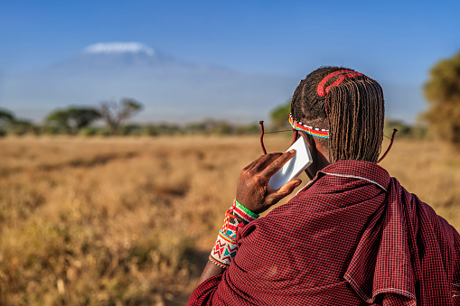 African warrior from Maasai tribe using mobile phone - Mount Kilimanjaro on the background, central Kenya, Africa. Maasai tribe inhabiting southern Kenya and northern Tanzania, and they are related to the Samburu.