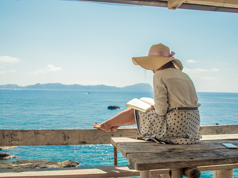 Young woman reading a book sitting on wooden balcony on Palmarola island in front of the ocean on a sunny day. Elegant white dress with skirt and hat.