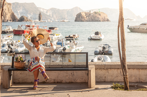 Young woman sitting on a bench at the beach. Ponza Island Harbour, Italy. Fashion dress, sunglasses, large hat. Boats in the background.
