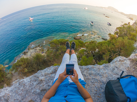 Young woman with backpack sitting on a cliff in front of the ocean using smartphone. Ponza Island coast, Italy. 8602