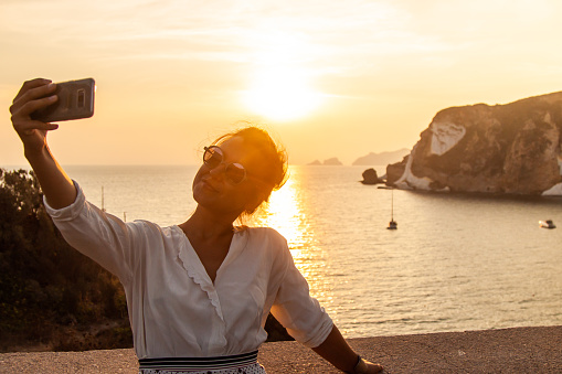 Young woman with smartphone taking selfies with sunset in front of the sea on Ponza island coast, on a wall with view of the ocean. 9811