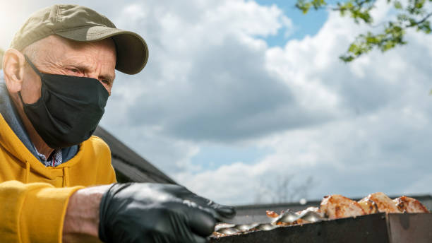 pensioner in black mask controls barbecue meat on grill