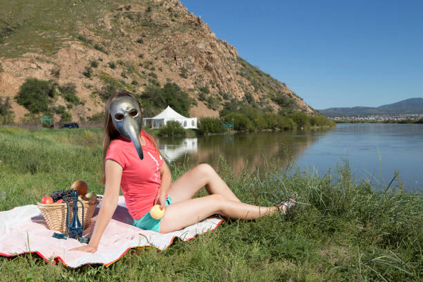 Young woman is relaxing on the lake in antiplague mask A young woman on a summer sunny day has a rest on the grassy shore of the lake in an anti-plague mask with fruits and drinks, enjoying the sun, wonderful nature, fresh air, sunbathing and having a picnic. black plague doctor stock pictures, royalty-free photos & images
