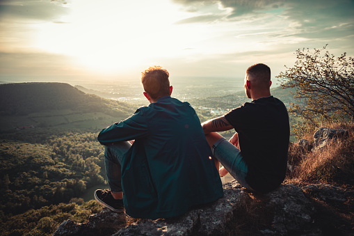 Best friends sitting together side by side on Mountain Top during sunset. Looking down to the valley, enjoying the sunset mood and view together. Millennial Generation Real People Outdoor Nature Lifestyle.