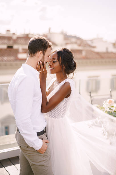 coppia di nozze di razza mista. sposo caucasico e sposa afro-americana coccole su un tetto alla luce del sole al tramonto. matrimonio d'arte a firenze. - couple sensuality embracing macho foto e immagini stock