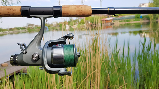 Fishing reel on a spinning comel close-up on the background of a lake or river. Reel with green fishing line on a natural background. Fisherman's equipment. Copy space. Natural background