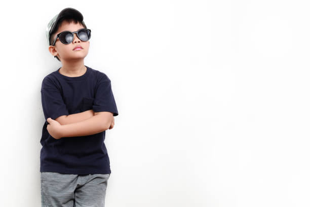Portrait of trendy Asian boy standing with arms crossed, wearing casual clothing, cap and sunglasses on white background.