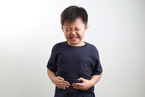 Little Asin boy holding aching stomach, standing against light background.