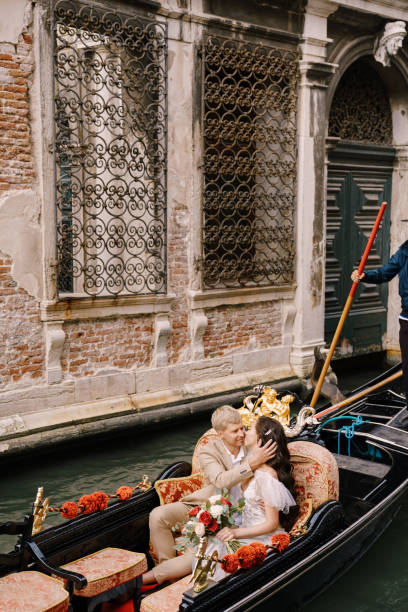 italien hochzeit in venedig. gondolier rollt braut und bräutigam in klassischer holzgondel entlang schmalen venezianischen kanal. newlyweds sitzen in boot nase zu nase, schwimmen vor dem hintergrund der alten geschmiedeten gitter - shoe groom wood luxury stock-fotos und bilder