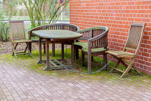A wooden seating group, consisting of a wooden table, wooden chairs and a wooden bench in front of a house wall, chained against theft and covered with a green layer of algae, moss and lichen, just like the paved floor.