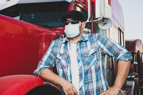 Semi truck professional driver on the job in casual clothing wears safety medical face mask. Confident looking trucker stands next to red big rig wearing protection sunglasses and surgical mask. conductor profesional de semi camión en el trabajo en ropa casual lleva máscara facial médica de seguridad. el camionero de aspecto seguro se encuentra junto a la gran plataforma roja que usa gafas de sol de protección y máscara quirúrgica.